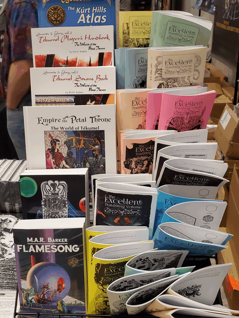 Display of Tekumel products for sale at NTRPG Con 2025. One side of the display shows the Tekumel Source Book, Empire of the Petal Throne, and two novels. The other side shows all thirteen issues of The Excellent Traveling Volume.